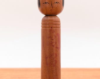 May include: A cylindrical wooden kokeshi doll with a natural brown wood grain finish. The doll features a rounded head with hand-painted black facial details, including arched eyebrows, closed eyes, and a small mouth, topped with a black hair detail.
