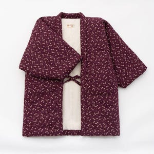 Könnte beinhalten: Eine gesteppte Hanten-Jacke in tiefem Pflaumenton mit einem kleinen weißen Blumenmuster. Das Kleidungsstück hat weite Ärmel, ein cremefarbenes Innenfutter und zwei dünne Stoffbänder in der vorderen Mitte zum Verschließen. Ein kleines Etikett ist sichtbar.