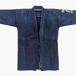 Peut inclure: Une veste bleu foncé teinte à l'indigo avec des coutures sashiko, des manches larges et une ouverture frontale. La manche gauche présente une broderie en kanji blancs, et le revers intérieur affiche des kanji rouges et une étiquette.