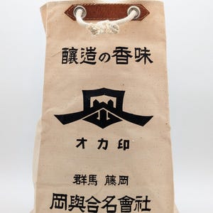 May include: A beige canvas drawstring bag featuring a brown leather patch at the top with silver metal grommets and a white rope cord. The bag displays black Japanese text and a central black graphic logo printed on the front surface.