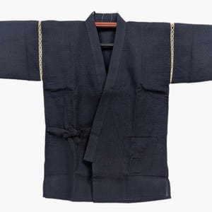 May include: A dark navy blue kimono-style jacket featuring wide sleeves with a decorative cream-colored braided trim along the shoulder seams. The garment has a wrap-front design with a tie closure at the waist and a single patch pocket on the front.