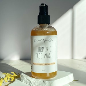 May include: A clear amber glass bottle with a black pump dispenser. The bottle is labeled "Oil and Honey Shop Turmeric Face Wash" and "Brightens + Gently Cleanses + Hydrates". The bottle is sitting on a white rock.
