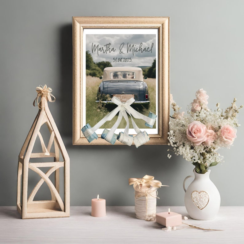 Charming wedding gift showcased in a wooden frame – a poster featuring wedding cars adorned with banknote-made cans. Frame put on gray wall. There are wooden, candle, vase, rose decorations on the desk under the frame.