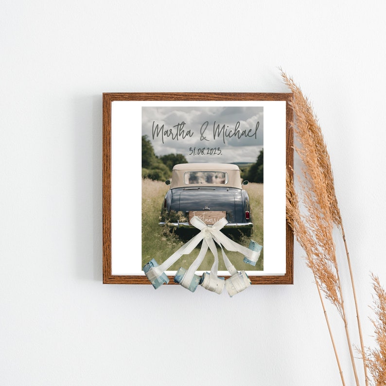 "Charming wedding gift showcased in a wooden frame – a poster featuring wedding cars adorned with banknote-made cans. Pampa grass decor on the right side of frame. Frame put on white background wall.