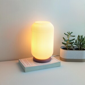 May include: A white ceramic table lamp with a ribbed design and a wooden base. The lamp is turned on and emitting a warm, yellow light. The lamp is sitting on a book on a white surface.