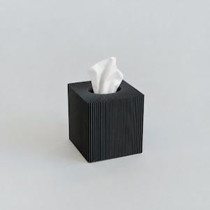 Pode incluir: Uma caixa quadrada preta para lenços com uma textura vertical canelada em todos os quatro lados. Um único lenço de papel branco é puxado através da abertura circular no centro do topo, sobressaindo da estrutura escura e estriada.