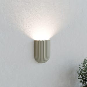 May include: A sage green wall sconce with a semi-circular, fluted design. The fixture is mounted to a textured white wall and emits a bright, upward-facing beam of light that illuminates the surface above the lamp in a soft, glowing arc.