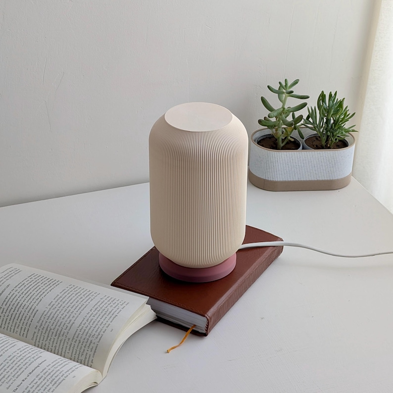 May include: A white ceramic table lamp with a ribbed design and a pink base. The lamp is sitting on a brown leather book on a white table. There are two small potted plants in the background.