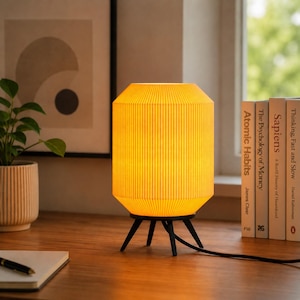 Puede incluir: Una lámpara de mesa amarilla con textura acanalada y base de trípode negra. La lámpara está encendida, emitiendo un brillo cálido. Libros y una planta en maceta son visibles en el fondo, sugiriendo un tema de decoración del hogar.