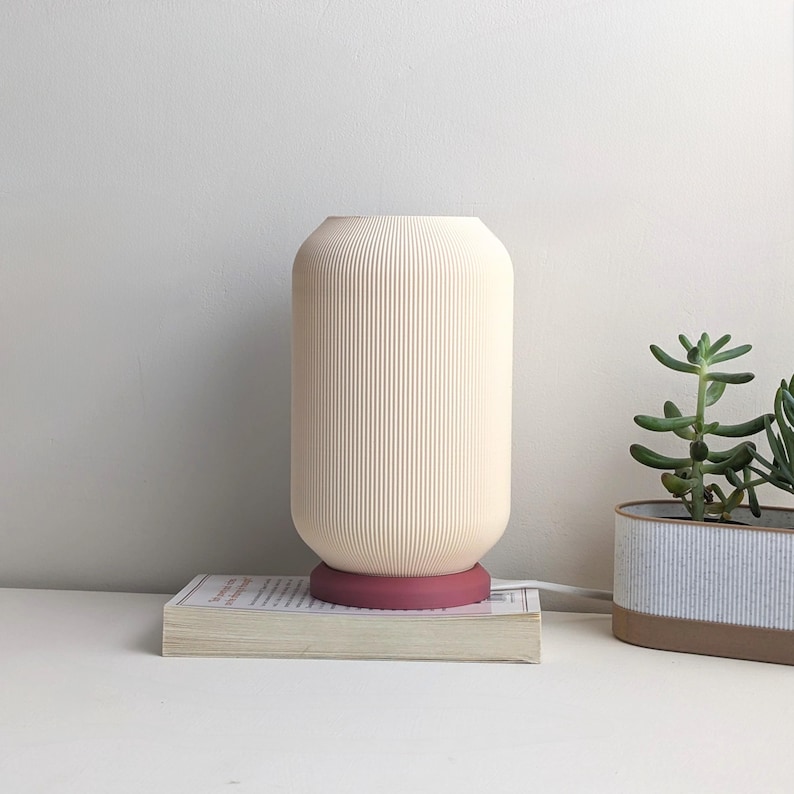 May include: A white ceramic table lamp with a ribbed design and a pink base. The lamp is sitting on a book on a white surface.