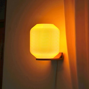 Puede incluir: Una moderna lámpara de pared cilíndrica con un cálido resplandor amarillo. La lámpara tiene un diseño acanalado y está montada sobre un soporte de madera. La luz ilumina la pared y una cortina cercana.