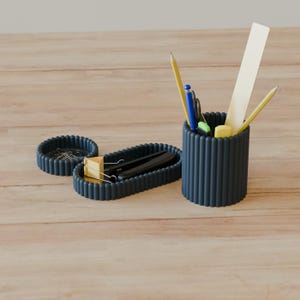 May include: A dark grey desk organiser set consisting of a cylindrical pen holder, an oval tray, and a small circular dish. The pen holder contains two yellow pencils, a blue pen, and a white ruler. The oval tray holds a black stapler.