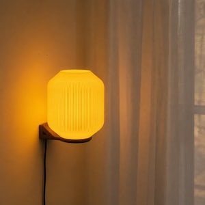 Op de afbeelding: Een wandlamp met een cilindervormige kap met verticale ribbels die een warm, geel licht uitstraalt. Het armatuur is met een donkere, hoekige beugel aan de muur bevestigd en is voorzien van een zwart stroomsnoer dat naar beneden loopt.