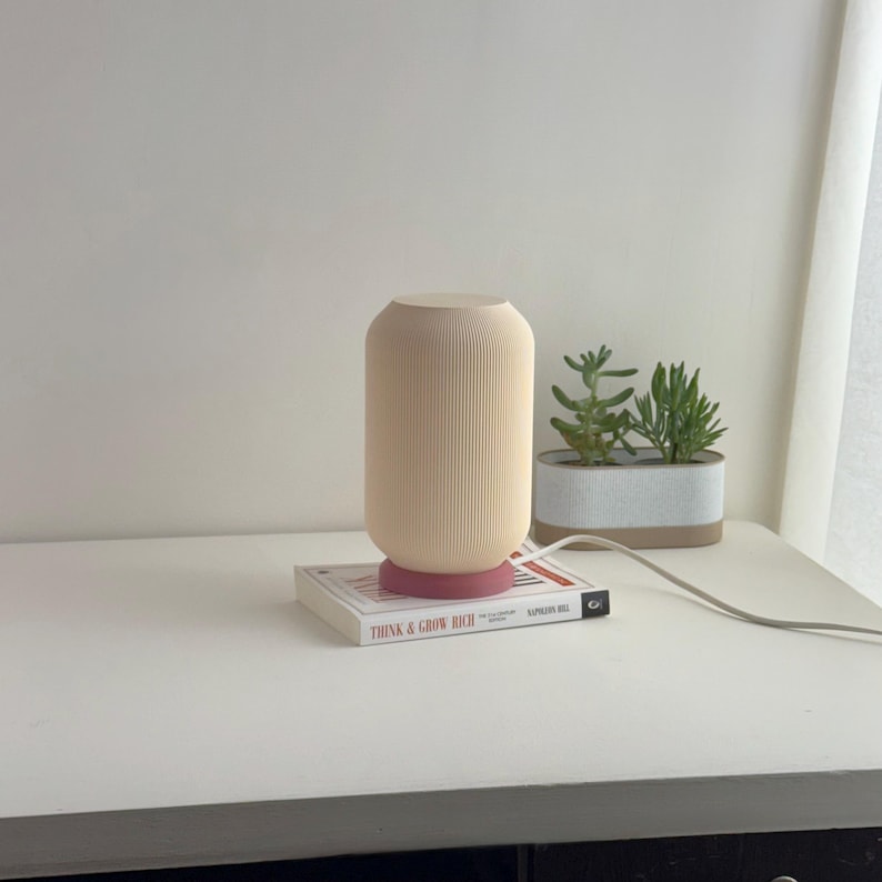 May include: A cream-colored table lamp with a ribbed design and a pink base sits on a book titled "Think & Grow Rich" by Napoleon Hill. The lamp is on a white desk with a potted succulent plant in the background.