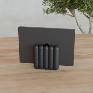 Puede incluir: Un soporte para tableta gris oscuro con cinco pilares verticales redondeados en forma de píldora dispuestos en fila. El soporte sostiene una tableta gris oscuro con un logotipo de Apple negro visible centrado en la parte posterior, colocada verticalmente sobre una superficie de madera clara.