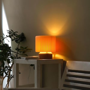 May include: A warm, orange-toned table lamp with a pleated fabric shade sits on a white side table. The lamp is turned on and casting a soft glow.