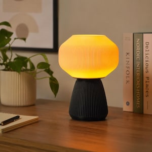 May include: A modern table lamp with a ribbed, yellow lampshade and a black base. The lamp is turned on, emitting a warm glow. The lamp is on a wooden surface, with books and a potted plant in the background. Home decor.