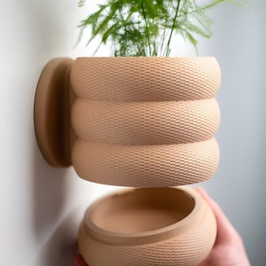 May include: A beige ceramic planter with a textured surface. The planter is designed to be mounted on a wall and has a removable top section. A green plant is growing inside the planter.