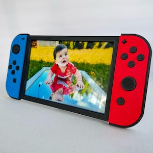 Peut inclure: Un cadre photo en forme de console Nintendo Switch. Le cadre est noir au centre, avec des côtés bleus et rouges imitant les manettes. Il contient une photo d'un bébé portant un t-shirt rouge.