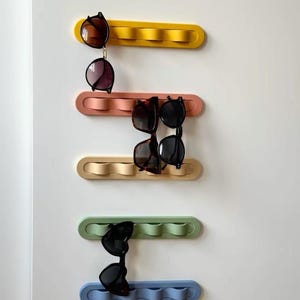 Peut inclure: Cinq organisateurs muraux horizontaux de couleur jaune, rose, beige, vert clair et bleu. Chaque organisateur présente un cadre rectangulaire arrondi avec trois crochets ondulés en saillie conçus pour suspendre des lunettes. Plusieurs paires de lunettes de soleil sont accrochées aux crochets, montrant diverses formes de montures et des verres foncés.