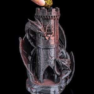 May include: A black and red dragon-themed dice tower with a gold and black die being placed on top. The tower has a castle design with dragon wings and a dragon wrapped around the tower. The dice tower is made of a dark material.