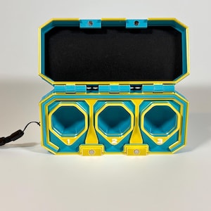 K&ouml;nnte beinhalten: Ein teal- und gelbfarbener Kunststoffbeh&auml;lter mit drei nummerierten F&auml;chern. Der Deckel ist ge&ouml;ffnet und gibt einen schwarzen Innenraum frei. Ein schwarzes Schl&uuml;sselband ist an der Seite befestigt. Der Beh&auml;lter ist mit geometrischen Formen und einer futuristischen &Auml;sthetik gestaltet.