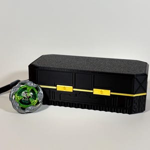 K&ouml;nnte beinhalten: Eine schwarze rechteckige Box mit goldenen Akzenten und einer strukturierten Oberseite, neben einem gr&uuml;nen und silbernen Spielzeug mit einem schwarzen Riemen. Das Spielzeug hat ein kreisf&ouml;rmiges Design mit gr&uuml;nen und silbernen Details.