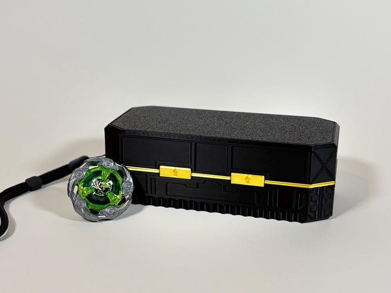 K&ouml;nnte beinhalten: Eine schwarze rechteckige Box mit goldenen Akzenten und einer strukturierten Oberseite, neben einem gr&uuml;nen und silbernen Spielzeug mit einem schwarzen Riemen. Das Spielzeug hat ein kreisf&ouml;rmiges Design mit gr&uuml;nen und silbernen Details.