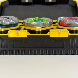 K&ouml;nnte beinhalten: Eine schwarze und gelbe Spielzeugaufbewahrungsbox mit drei bunten Kreiselspielzeugen. Die Kreisel sind silber, gr&uuml;n und rot und stehen auf gelben Plattformen. Die Box hat einen gelben Deckel und eine schwarze Basis mit strukturiertem Design.