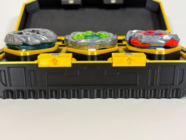 K&ouml;nnte beinhalten: Eine schwarze und gelbe Spielzeugaufbewahrungsbox mit drei bunten Kreiselspielzeugen. Die Kreisel sind silber, gr&uuml;n und rot und stehen auf gelben Plattformen. Die Box hat einen gelben Deckel und eine schwarze Basis mit strukturiertem Design.