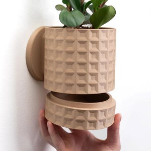 May include: A beige ceramic planter with a geometric pattern. The planter is designed to be mounted on a wall and has a removable bottom section.