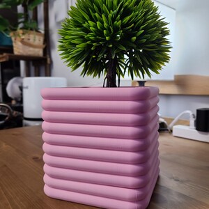 K&ouml;nnte beinhalten: Ein quadratischer, rosafarbener Pflanzer mit abgerundeten Kanten und einem gerippten Design. Der Pflanzer ist mit einem gr&uuml;nen, k&uuml;nstlichen Busch gef&uuml;llt.
