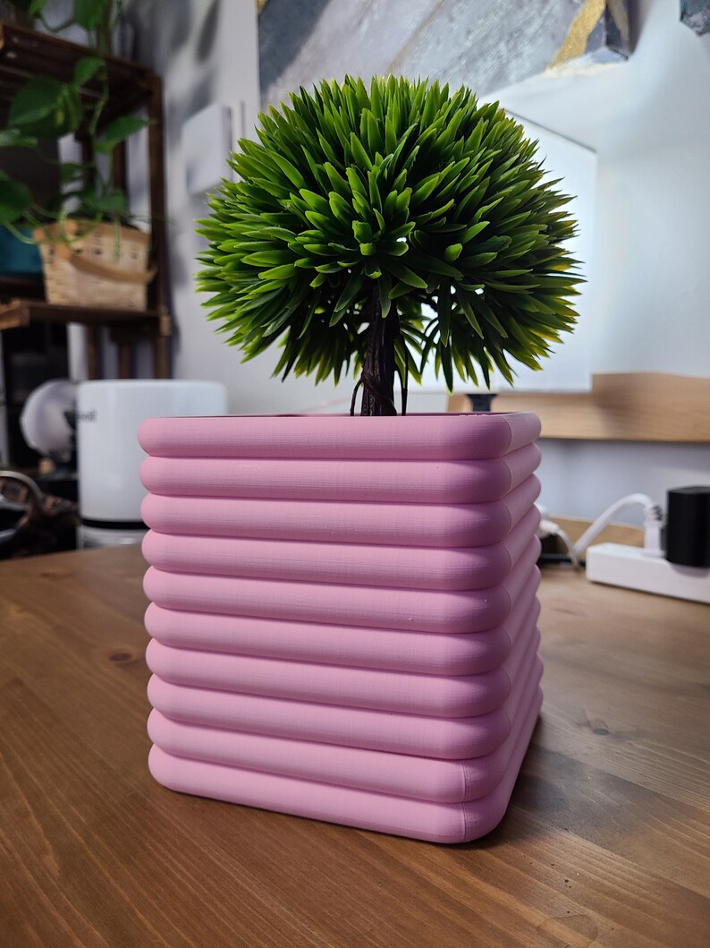 K&ouml;nnte beinhalten: Ein quadratischer, rosafarbener Pflanzer mit abgerundeten Kanten und einem gerippten Design. Der Pflanzer ist mit einem gr&uuml;nen, k&uuml;nstlichen Busch gef&uuml;llt.