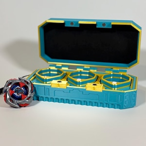 K&ouml;nnte beinhalten: Eine teal- und goldfarbene Spielzeugaufbewahrungsbox mit drei F&auml;chern, die sich &ouml;ffnet, um einen schwarzen Innenraum freizulegen. Ein rotes, silbernes und blaues Spielzeug befindet sich neben der Box. Die Box hat eine goldfarbene Umrandung und einen passenden goldenen Verschluss.