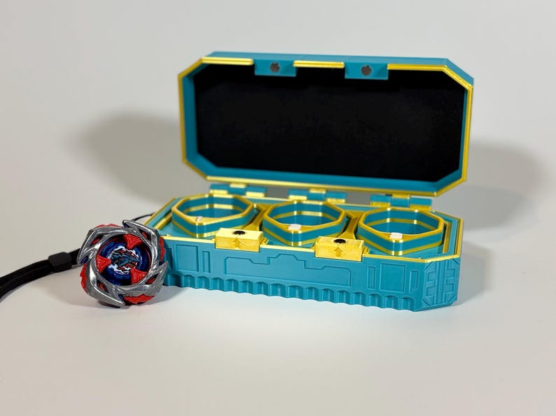 K&ouml;nnte beinhalten: Eine teal- und goldfarbene Spielzeugaufbewahrungsbox mit drei F&auml;chern, die sich &ouml;ffnet, um einen schwarzen Innenraum freizulegen. Ein rotes, silbernes und blaues Spielzeug befindet sich neben der Box. Die Box hat eine goldfarbene Umrandung und einen passenden goldenen Verschluss.