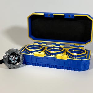 K&ouml;nnte beinhalten: Eine blaue und goldene Aufbewahrungsbox mit drei F&auml;chern f&uuml;r Kreisel. Ein grauer und schwarzer Kreisel mit einem Wolfsemblem befindet sich neben der Box. Die Box ist ge&ouml;ffnet und zeigt einen schwarzen Innenraum.
