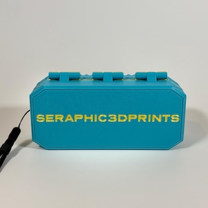 K&ouml;nnte beinhalten: Eine t&uuml;rkisblaue 3D-gedruckte Box mit einem schwarzen Schl&uuml;sselband. Die Box hat eine rechteckige Form mit einer Reihe von erhabenen rechteckigen Merkmalen auf der Oberseite. Der Text "SERAPHIC3DPRINTS" ist goldfarben auf die Vorderseite der Box gedruckt.