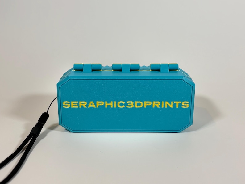 K&ouml;nnte beinhalten: Eine t&uuml;rkisblaue 3D-gedruckte Box mit einem schwarzen Schl&uuml;sselband. Die Box hat eine rechteckige Form mit einer Reihe von erhabenen rechteckigen Merkmalen auf der Oberseite. Der Text "SERAPHIC3DPRINTS" ist goldfarben auf die Vorderseite der Box gedruckt.