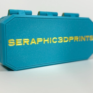 K&ouml;nnte beinhalten: Ein t&uuml;rkisfarbenes, rechteckiges Objekt mit goldfarbenem Schriftzug "SERAPHIC3DPRINTS". Das Objekt hat eine strukturierte Oberfl&auml;che und einige erhabene rechteckige Merkmale auf der Oberseite. Die Kanten sind goldfarben akzentuiert.