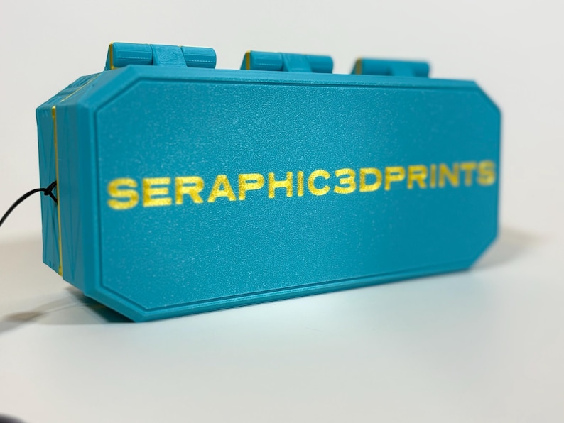 K&ouml;nnte beinhalten: Ein t&uuml;rkisfarbenes, rechteckiges Objekt mit goldfarbenem Schriftzug "SERAPHIC3DPRINTS". Das Objekt hat eine strukturierte Oberfl&auml;che und einige erhabene rechteckige Merkmale auf der Oberseite. Die Kanten sind goldfarben akzentuiert.