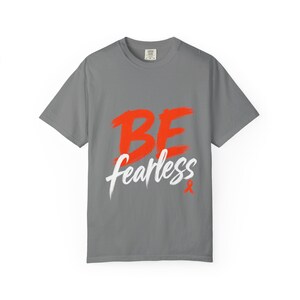 May include: A gray t-shirt with the words "BE fearless" in a bold orange and white design. The shirt has a crew neck and short sleeves. A small orange ribbon is at the bottom.