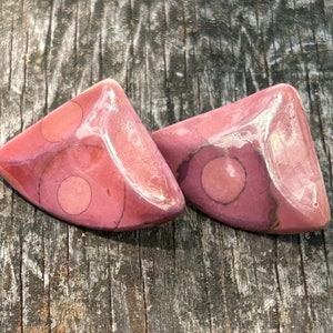 Puede incluir: Un par de pendientes triangulares de color rosa con círculos blancos y rosas. Los pendientes están hechos de un material liso y pulido.