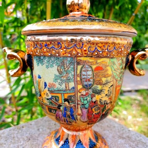 May include: A porcelain vase with a lid, featuring a gold trim and intricate hand-painted scenes of people and landscapes in vibrant colors. The vase has a round body and a pedestal base.