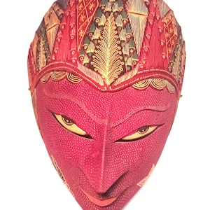 Può includere: Una maschera di legno rossa con intricati motivi dorati e verdi. La maschera ha un'espressione sorridente con occhi a mandorla.