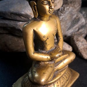 May include: A small, brass statue of a seated Buddha. The statue is highly detailed, with intricate carvings on the base and clothing. The Buddha's face is serene and peaceful, and the overall color is a rich, golden brass. The statue is a collectible item, perfect for meditation or display.