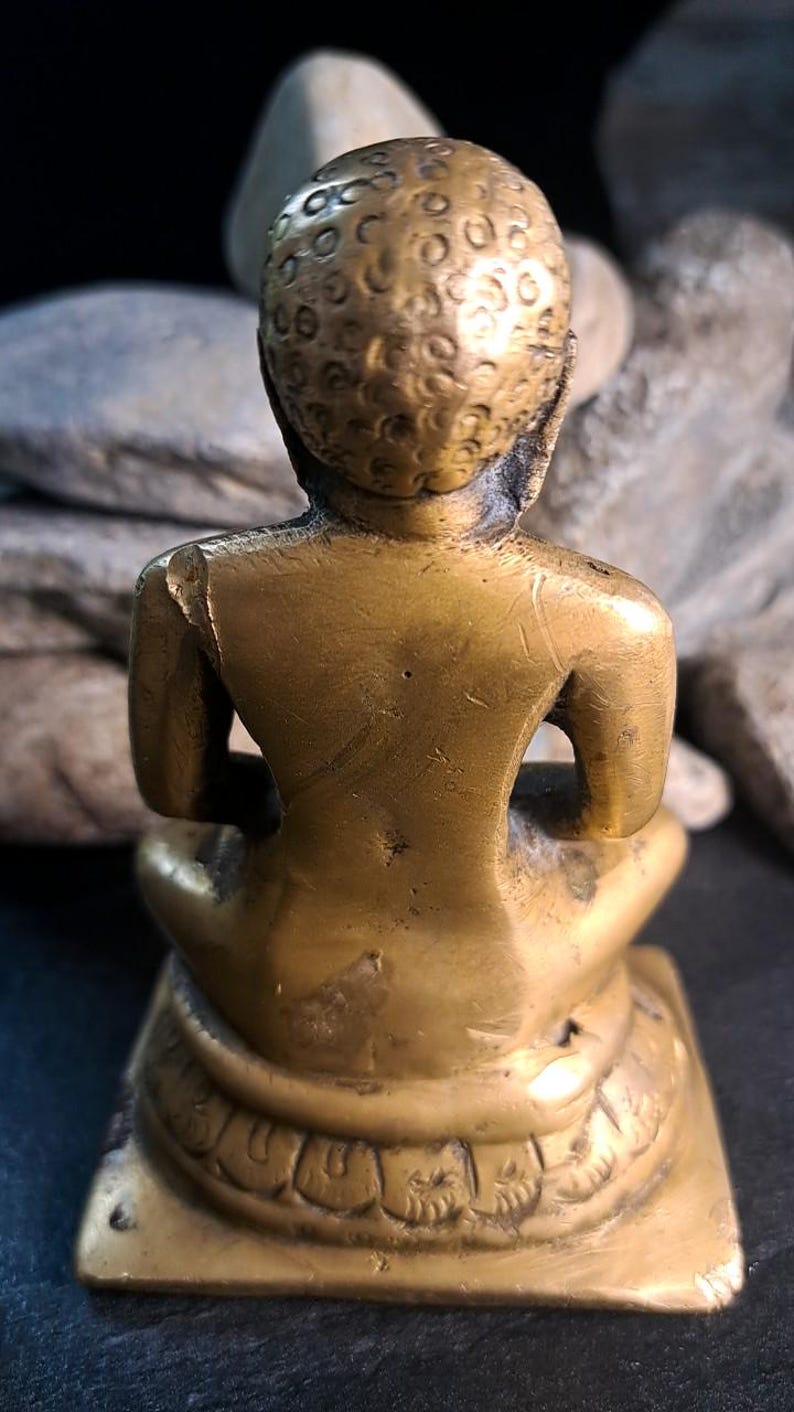 May include: A brass statue of a person in a seated meditation pose. The statue is gold-colored and has intricate details on the head and base. The back of the statue shows some minor damage.  The statue is a religious figurine, possibly depicting a Buddha or similar figure. The base is square and features decorative carvings. The overall style suggests an antique or vintage piece.