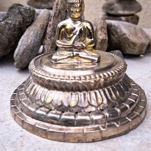 May include: A gold-colored Buddha statue sitting in meditation pose on a silver base with intricate details. The base has a lotus flower design.