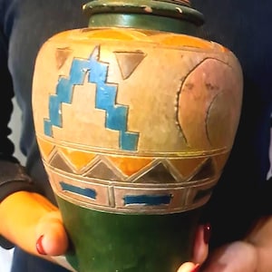 May include: A decorative ceramic jar with a lid. The jar features a green base, a tan body with geometric blue, orange, and brown designs, and a green and orange lid. The jar is held by a person.