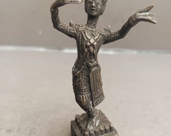 Może przedstawiać: Mała brązowa statuetka tancerki z rozciągniętymi ramionami, ubrana w tradycyjny strój i koronę. Statuetka stoi na kwadratowej podstawie.