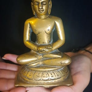May include: A brass statue of a seated figure, possibly a deity or religious figure, in a meditative pose. The statue has a serene expression and intricate details. The figure is adorned with a headdress and sits on a lotus-shaped base. The statue is golden in color with a slightly aged patina, suggesting it may be antique or vintage. The overall style is traditional and possibly of Asian origin.